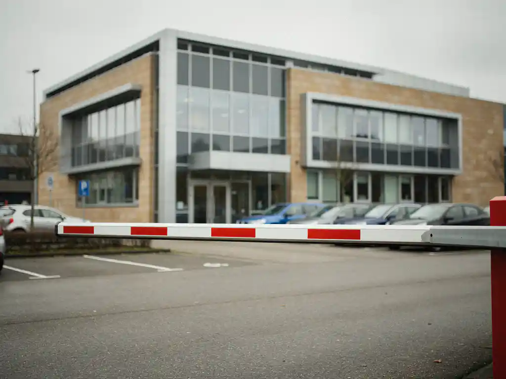 Gestreepte parkeerslagboom voor een modern glazen kantoorpand met bijna vol parkeerterrein onder bewolkte Nederlandse lucht.